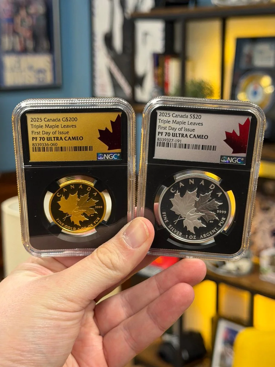 A hand is holding two collectible coins encased in protective holders. The left coin is gold, labeled "2023 Canada $5.00 Triple Maple Leaves – First Day of Issue PF 70 Ultra Cameo." The right coin is silver, labeled "2023 Canada $20 Triple Maple Leaves – First Day of Issue PF 70 Ultra Cameo." Both feature maple leaf designs. Background includes blurred indoor decor.