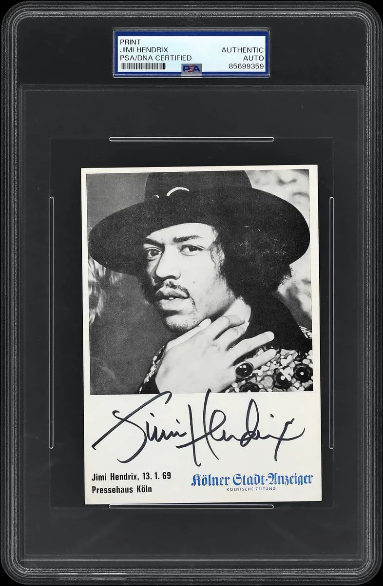 A signed black and white photograph of a person wearing a wide-brimmed hat and patterned clothing. The signature reads "Jimi Hendrix." The photo is encased in a PSA/DNA certified protective frame with the code 85601836. Additional text includes "Jimi Hendrix, 13. 1. 69, Pressehaus Köln" and "Kölner Stadt-Anzeiger.