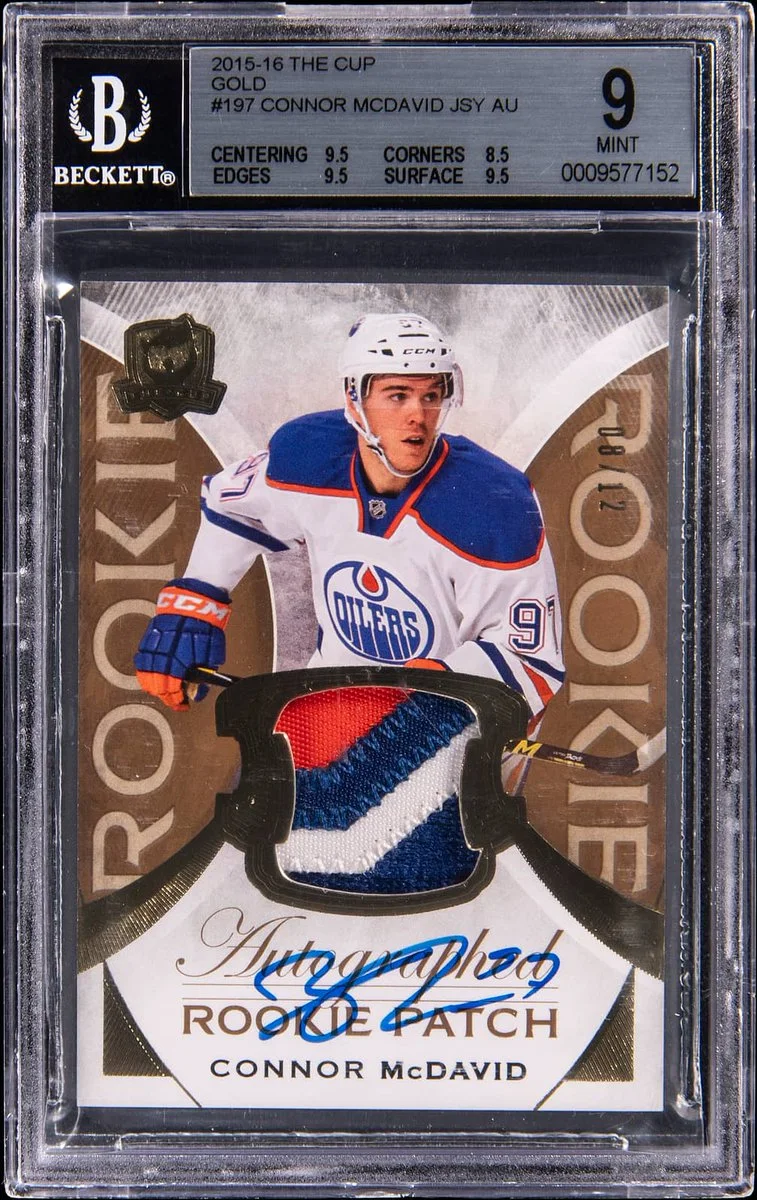 A graded Beckett hockey trading card of Connor McDavid, featuring a photo of the player in an Edmonton Oilers jersey. The card includes a piece of the jersey and is inscribed with "ROOKIE PATCH" and Connor McDavid's signature. It is labeled as "2015-16 The Cup Gold" and has a mint grade of 9.