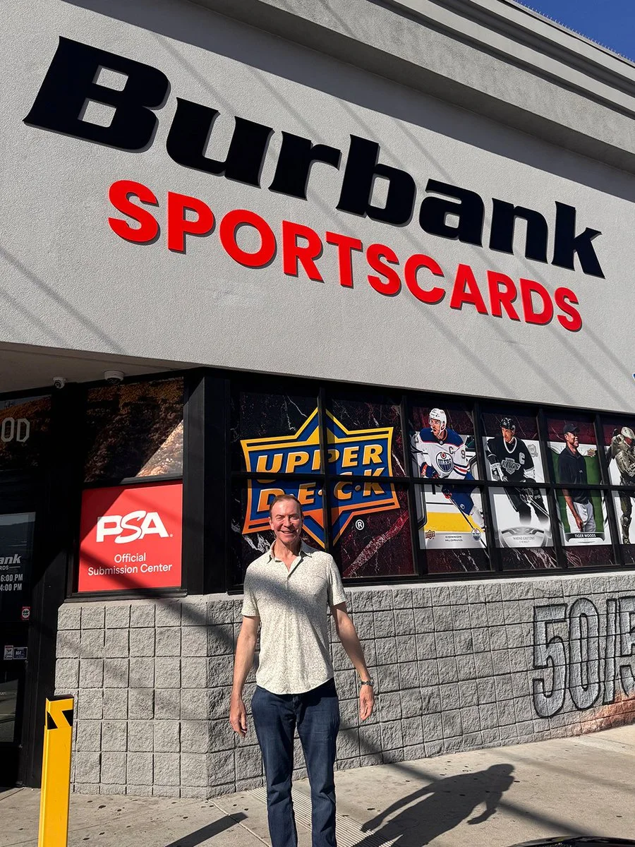 A person stands smiling in front of Burbank Sportscards, a store with large signage. The window displays logos of PSA and Upper Deck, along with images of sports figures.