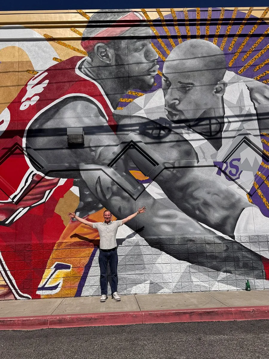 A person stands with arms outstretched in front of a large mural featuring two basketball players in action. One player is wearing a red jersey and the other a white one. Various colors and dynamic lines emphasize their movement on the mural.