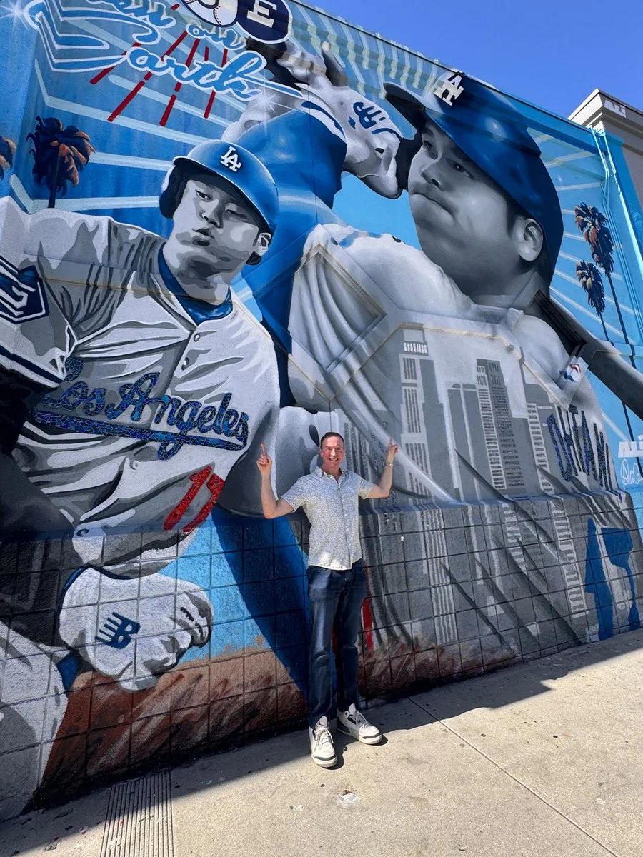 A person stands in front of a vibrant mural featuring baseball players in Los Angeles Dodgers uniforms. The artwork highlights dramatic action poses and includes the Los Angeles skyline in the background. The person points upwards toward the mural.