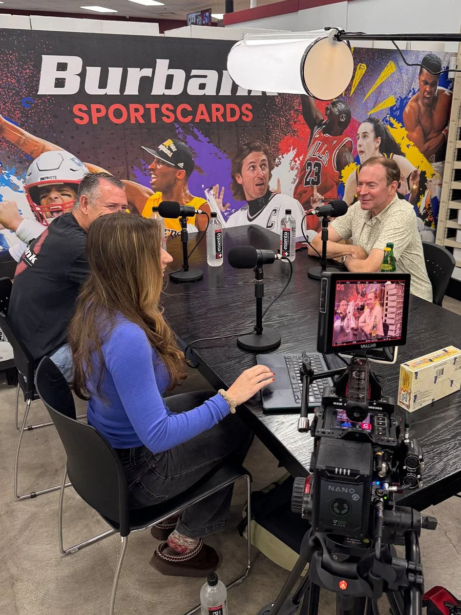 Four people are sitting around a black table with microphones, recording in a studio decorated with sports-themed posters. A camera on a tripod is set up in front of the table, showing a view of the participants. Burbank Sportscards signage is visible in the background.