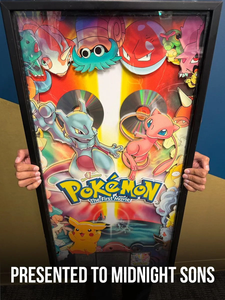 A person is holding a framed poster for "Pokémon: The First Movie" featuring various Pokémon characters such as Mewtwo, Mew, Pikachu, and others. The poster is vibrant and colorful, with the words "Presented to Midnight Sons" at the bottom.