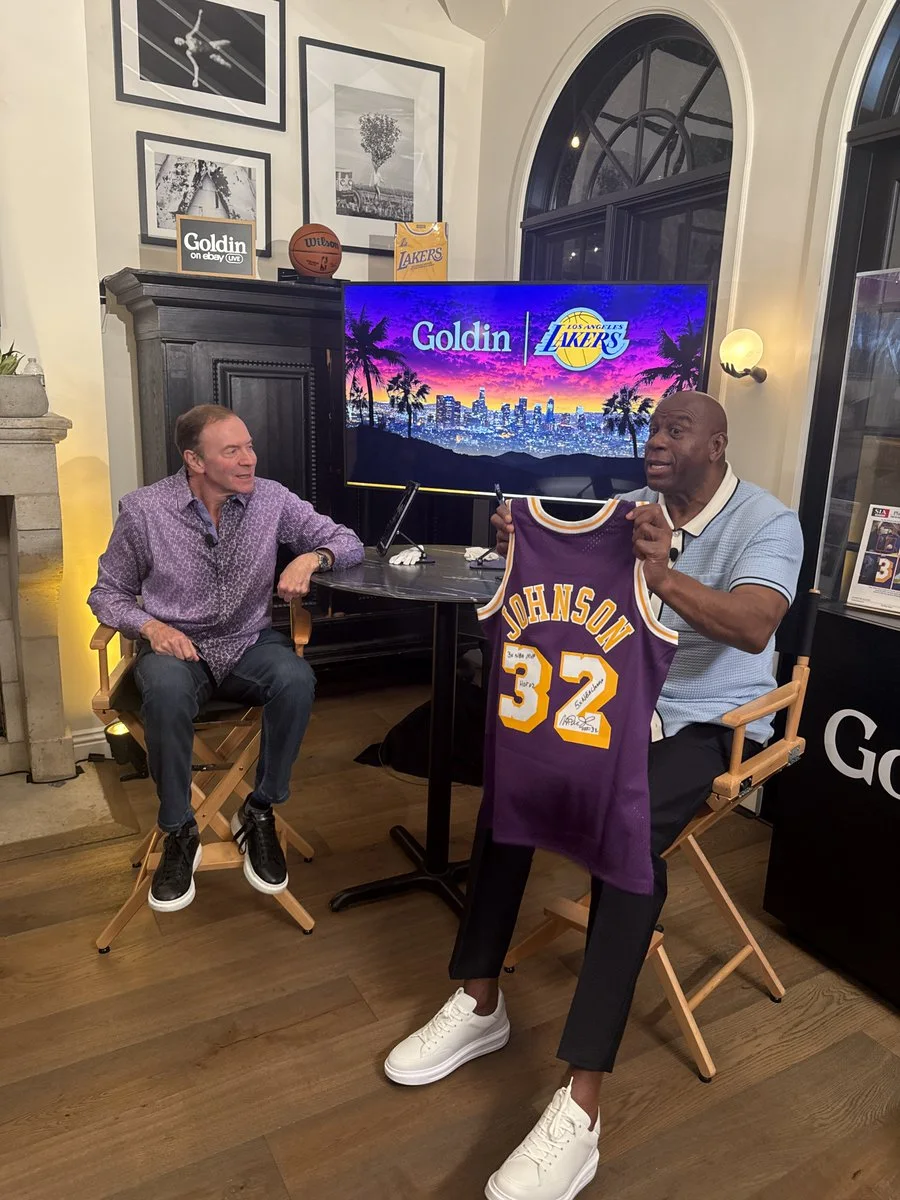 Two people are sitting indoors during a live stream or interview setting. One is holding a Los Angeles Lakers jersey with the number 32 and the name "Johnson." Behind them is a monitor displaying "Goldin" and a Lakers logo. Basketball-themed decor is visible, including a ball and related artwork.