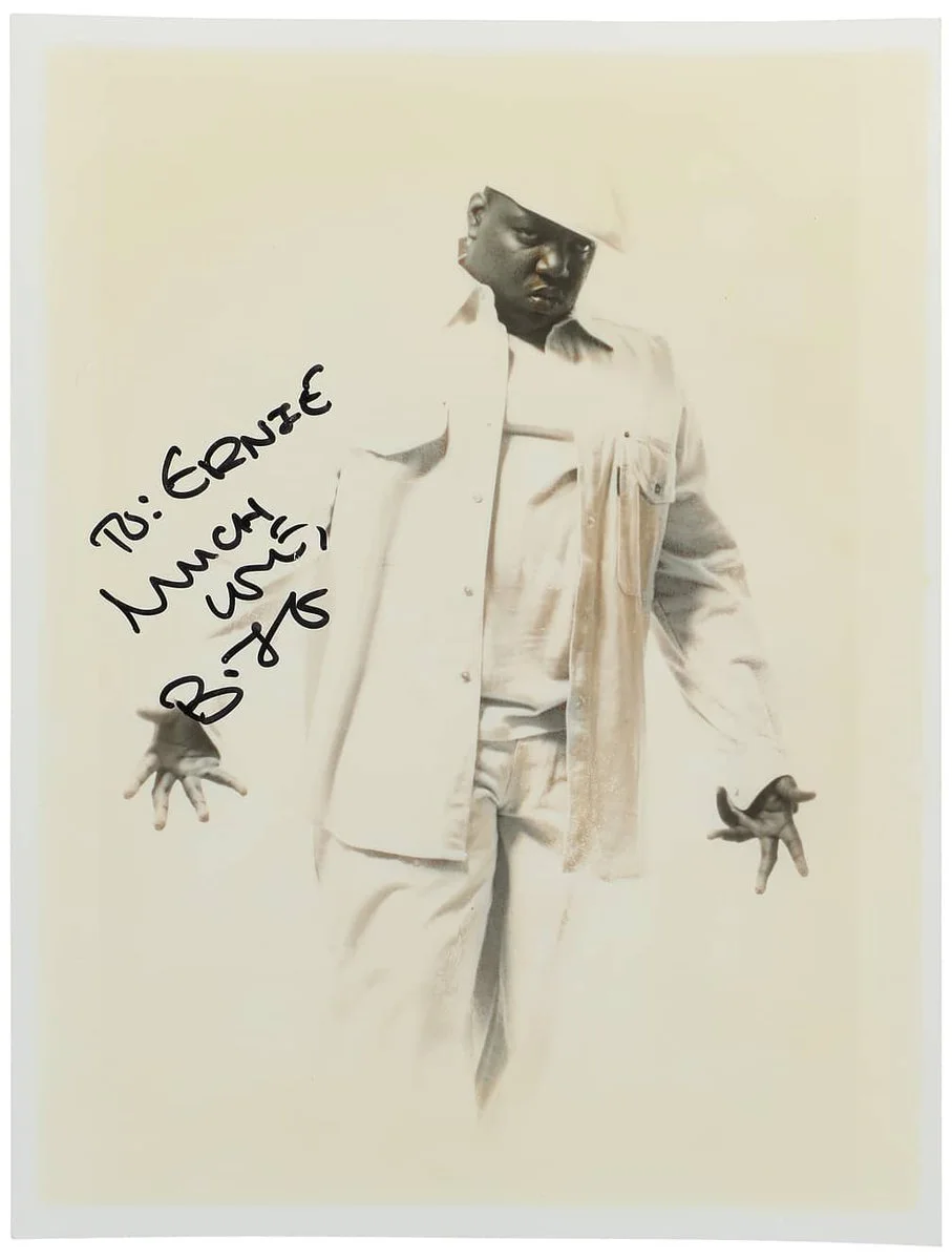 A person dressed in an all-white outfit, including a hat, poses with hands gesturing downward. A handwritten note on the left reads, βTo: Ernie, Much Love, Biggie.β