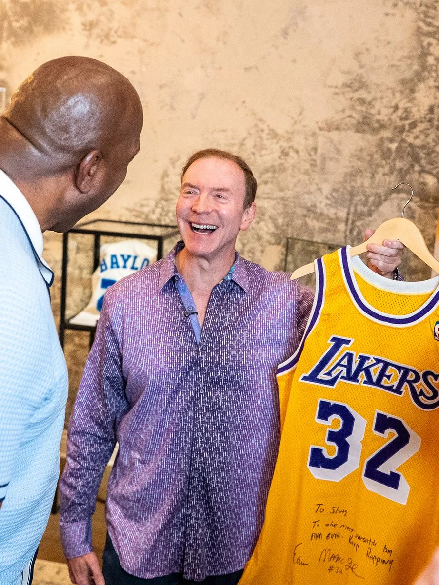 A person holds a Los Angeles Lakers jersey with the number 32 and a handwritten message. Another person stands nearby, facing the jersey holder. A jersey in a frame with the word "Baylor" is visible on the wall in the background.