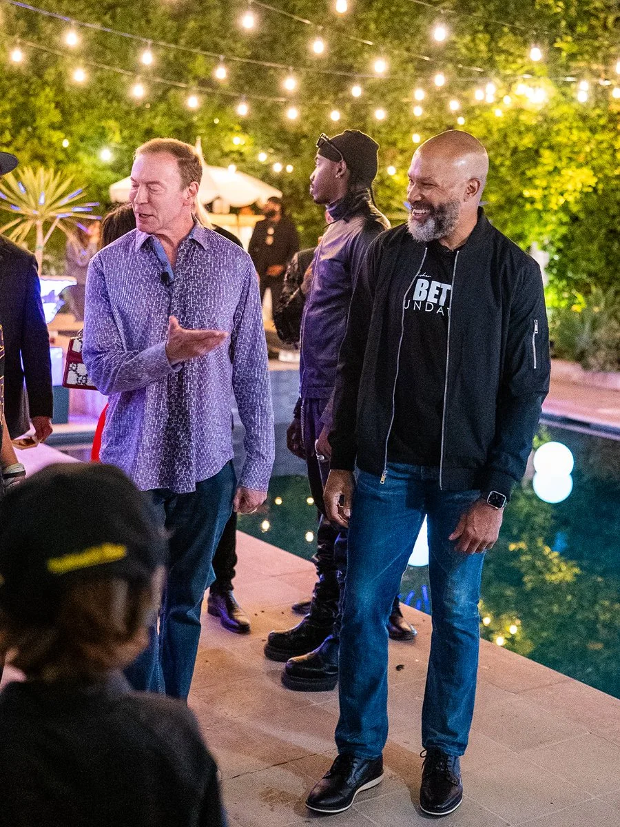A social gathering by a pool with several people interacting. Two individuals are engaged in conversation; one is wearing a patterned shirt, the other a black jacket and jeans. In the background, there are decorative lights strung above, and trees are visible, creating a festive atmosphere.