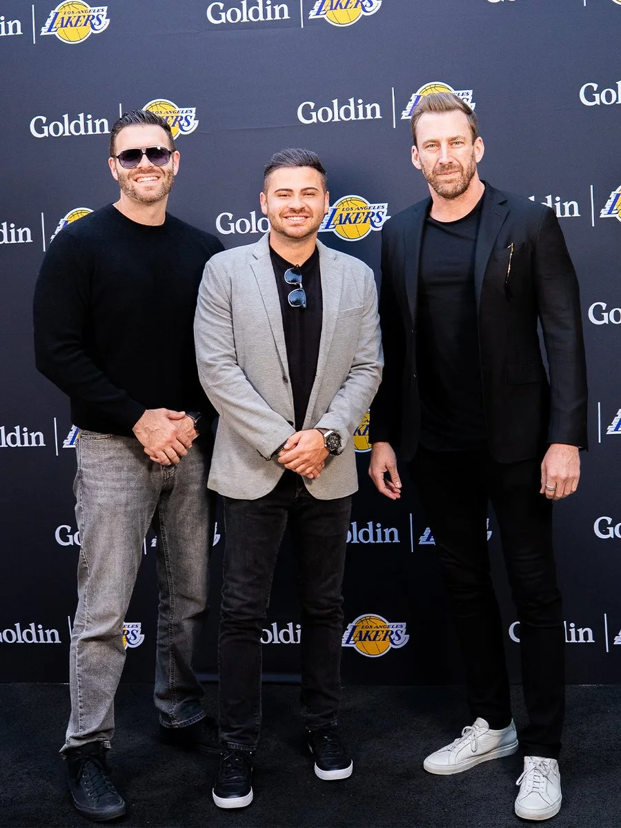 Three people stand in front of a backdrop featuring "Goldin" and Los Angeles Lakers logos. They are dressed in casual and business attire, smiling for the photo.