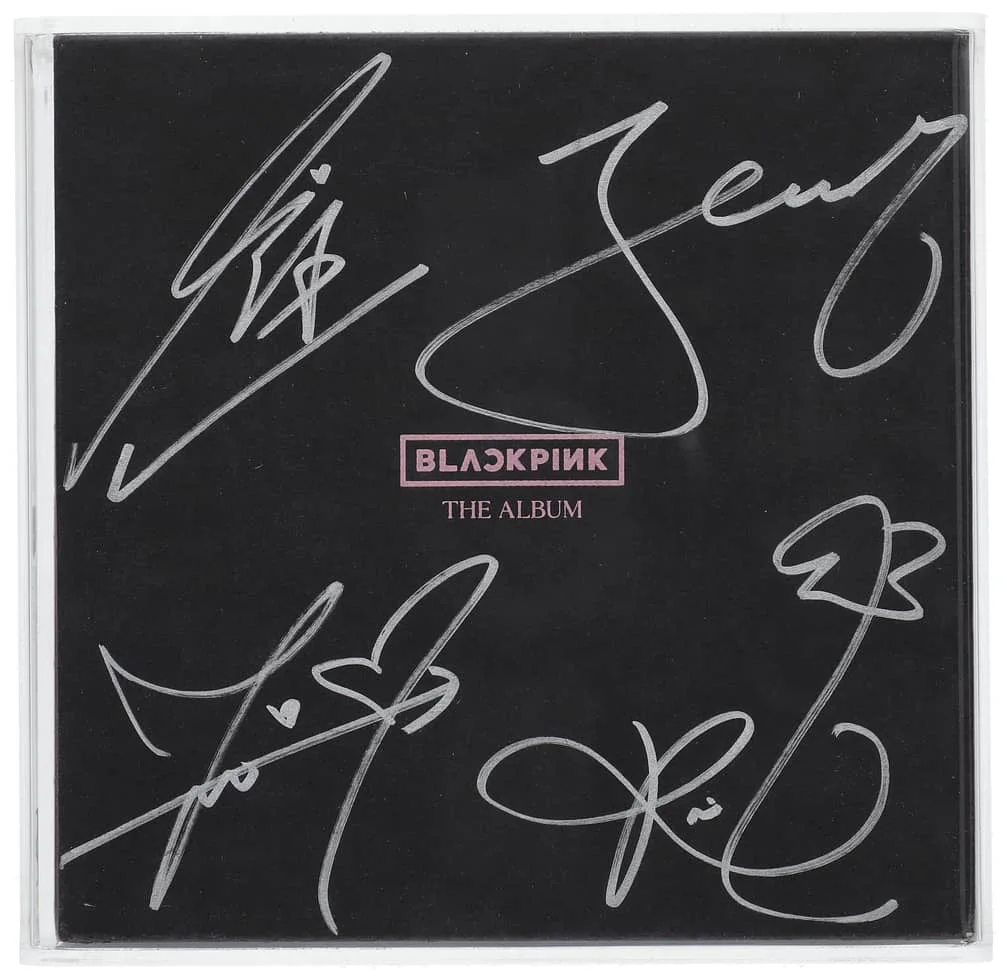 A BLACKPINK album cover with signatures in silver ink. The cover is predominantly black with the band's name and "THE ALBUM" in pink and white text at the center.