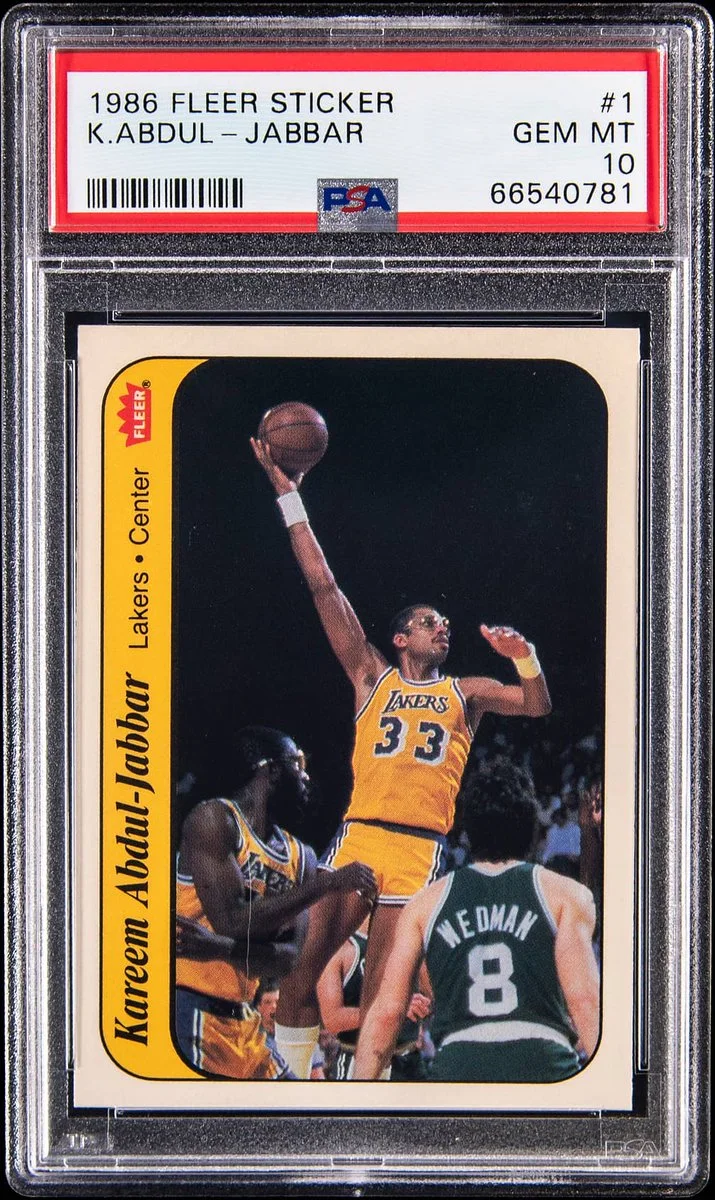A 1986 Fleer Sticker trading card graded by PSA as GEM MT 10. The card features Kareem Abdul-Jabbar in a Lakers uniform, jumping with a basketball. A Celtics player is in the background. The card displays the name, position, and team of the player.