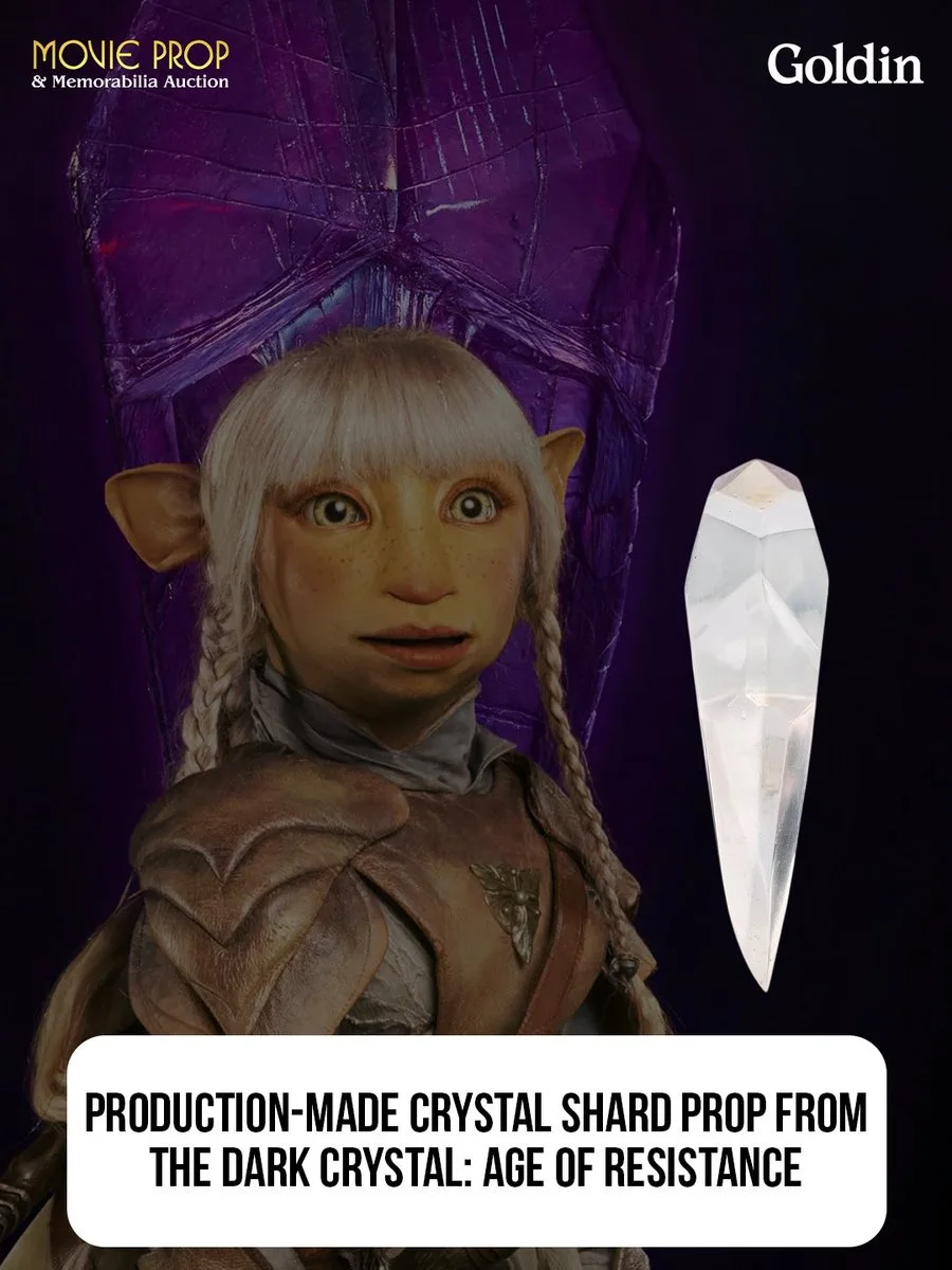 This image shows a character from "The Dark Crystal: Age of Resistance" standing next to a large purple crystal. In the foreground, there's a crystal shard prop. Text at the top indicates a movie prop auction by Goldin. At the bottom, there's a description of the crystal shard.