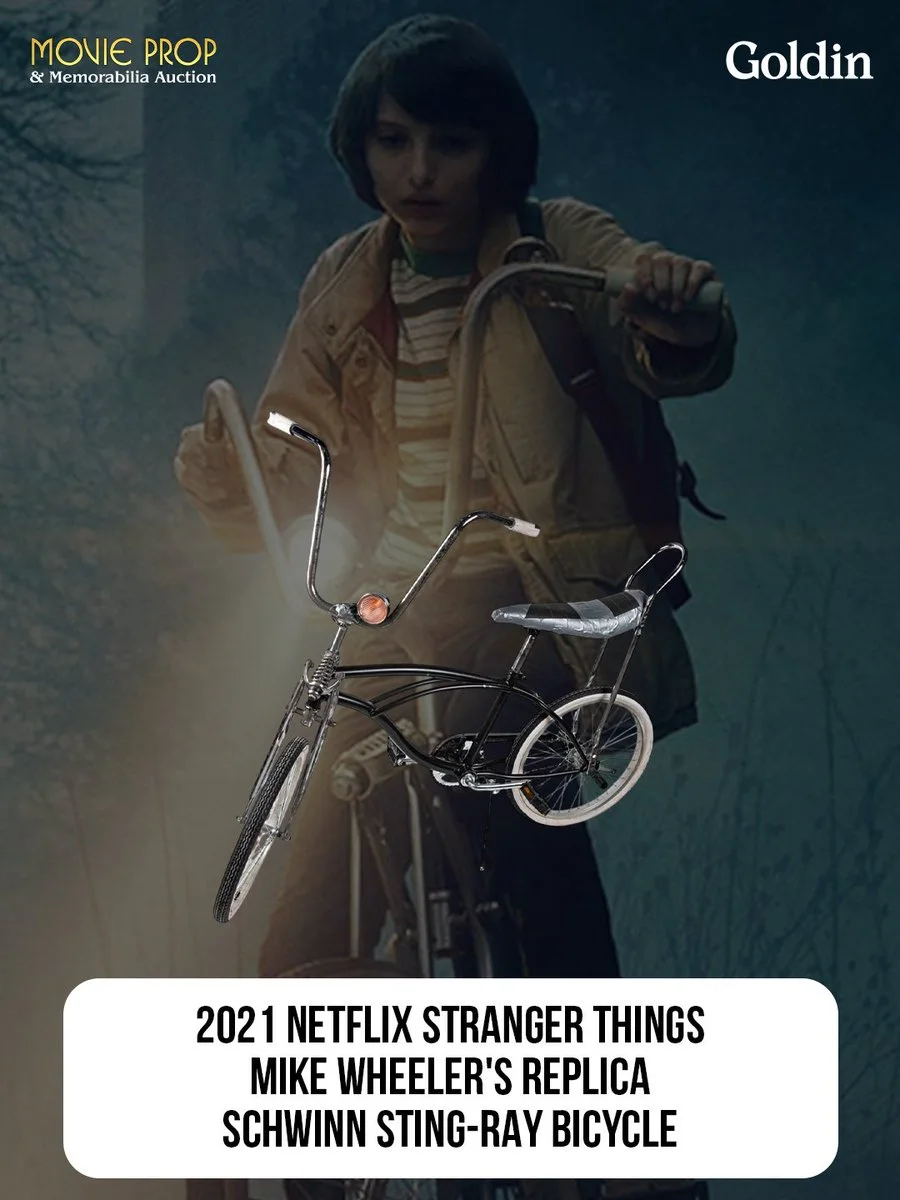A person rides a bicycle in a scene from "Stranger Things," with the bicycle highlighted in the foreground. The text highlights it as a replica of Mike Wheeler's Schwinn Sting-Ray bicycle from the 2021 Netflix series. Logos for Movie Prop & Memorabilia Auction and Goldin are at the top.
