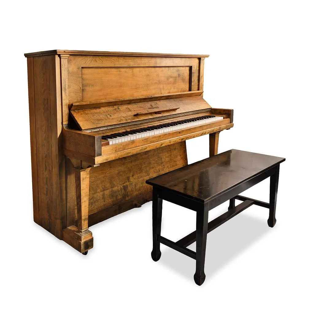 A wooden upright piano is positioned next to a dark wooden bench on a white background. The piano has a classic design with exposed keys.
