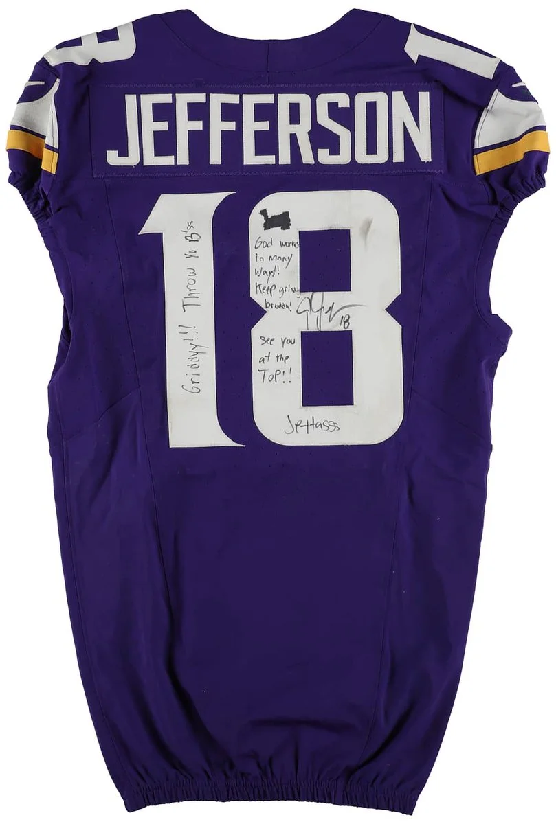 A purple football jersey with the number 18 and the name "JEFFERSON" on the back. The jersey is signed with messages that include "Griddy!!!” and “God works in crazy ways! Hope you know that!” The text is signed with “J. Jettas.”