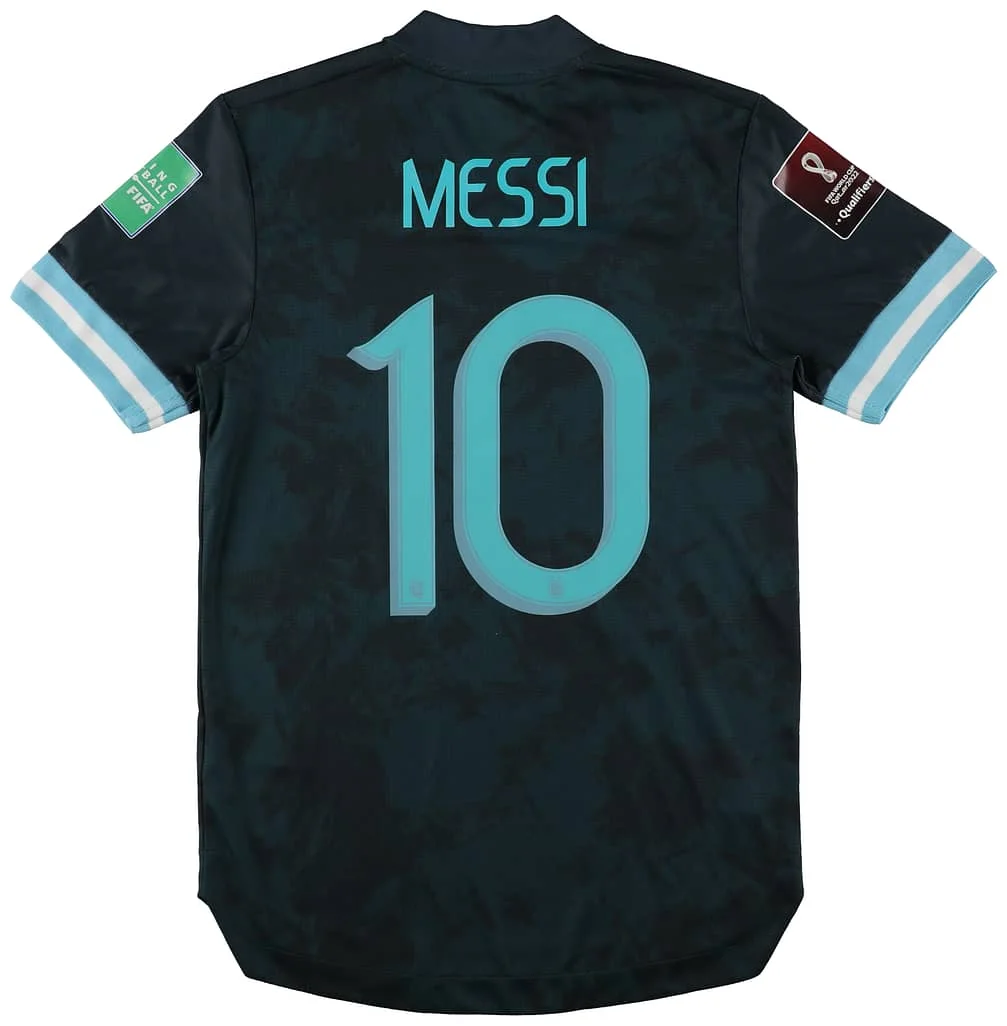 A dark sports jersey with "Messi" and the number 10 on the back in light blue. The sleeves feature light blue and white stripes, and there are event patches on both sleeves.