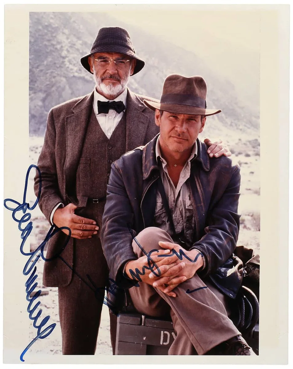 Two individuals dressed in 1930s-style adventure clothing are posing outdoors in a rocky, rural setting. One is standing, wearing a tweed suit, bow tie, and a felt hat. The other is sitting with legs crossed, wearing a leather jacket and a fedora. Both have signatures on the image.