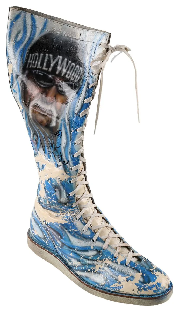 A tall, lace-up boot featuring a painted design. The artwork includes an image of a face wearing sunglasses, with the word "HOLLYWOOD" on a cap. The boot is adorned with blue flames and splattered white paint.